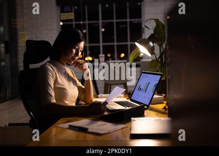 Happy asian businesswoman at desk, reading documents and using laptop, working late at office Stock Photo
