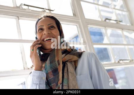 Happy biracial muslim biracial businesswoman in headscarf talking on smartphone in modern office Stock Photo