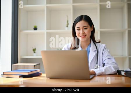 Attractive and smart millennial Asian female doctor in a uniform using her laptop at her desk in her office. Stock Photo