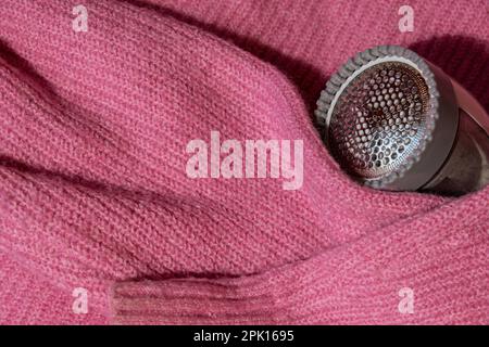 Anti-pilling razor. Device for shaving clothes. Anti-Plush fabric Shaver. Electric portable sweater pill defuzzer Lint remover from pink acrylic or wool sweater. Electric device fabric shaver after cleaning and collected fluff lint. Closeup Pilled sweater. Old used clothes with pilling.  Stock Photo