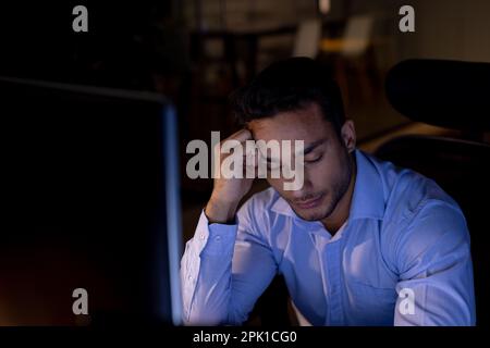 Tired biracial businessman sitting at desk and using computer, working late at office Stock Photo