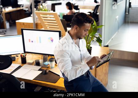 Biracial businessman leaning on desk and using tablet, working at office Stock Photo