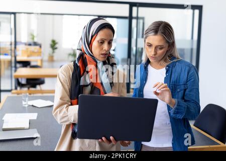Focused diverse casual businesswomen discussing work and using laptop in modern office Stock Photo