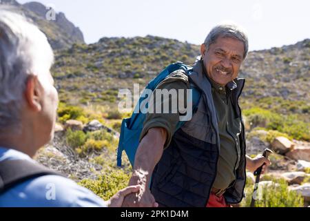 Happy senior biracial couple wearing backpacks, holding hands and hiking with trekking poles Stock Photo