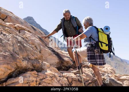 Happy senior biracial couple wearing backpacks, holding hands and hiking with trekking poles Stock Photo