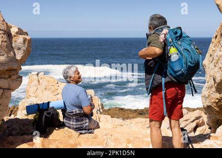 Happy senior biracial couple sitting on rocks by the sea Stock Photo