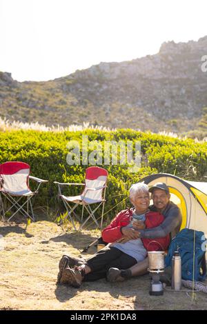 Happy senior biracial couple sitting in tent, embracing and drinking coffee in mountains Stock Photo