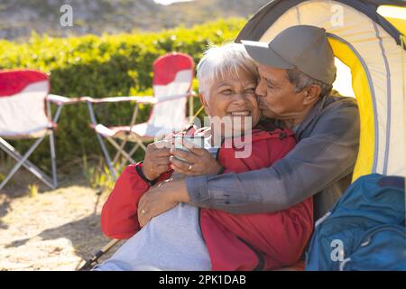Happy senior biracial couple sitting in tent, embracing and drinking coffee in mountains Stock Photo