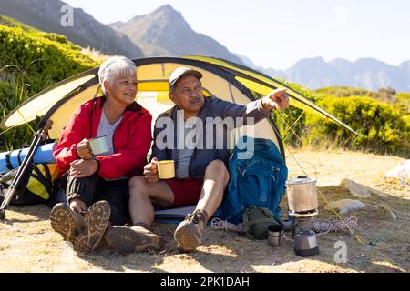 Happy senior biracial couple sitting in tent and drinking coffee in mountains Stock Photo