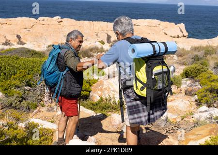 Senior biracial couple wearing backpacks, hiking with trekking poles Stock Photo
