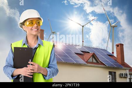 Industrial engineer in uniform and view of wind energy turbines near house with installed solar panels on roof Stock Photo