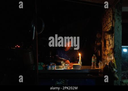 A vendor makes tea by candlelight during a blackout at a shop on the street in Yangon. Due to soaring costs and the lack of hydropower during the dry season, the military junta (Tatmadaw) rations electricity throughout Yangon daily, leaving large sections of the city (townships) completely blacked out everyday. Daily life during the deadly civil war in Myanmar. On February 1, 2021, the military junta government (Tatmadaw) seized power by coup, jailing the democratically-elected government and plunging the country into an ongoing humanitarian crisis. Stock Photo