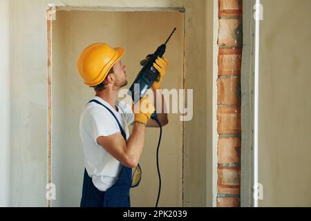 Holding electric drill in hands. Young man working in uniform at construction at daytime Stock Photo