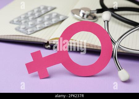 Female gender sign near open notebook, stethoscope and blisters of pills on violet background, closeup. Women's health concept Stock Photo