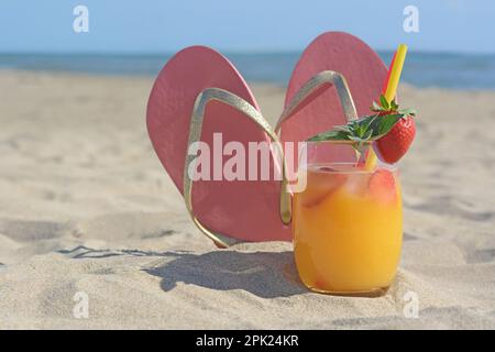 Glass of refreshing drink with strawberry and flip flops on sandy beach near sea Stock Photo