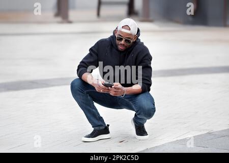 Cool African American man using smartphone, squatting on city street, wears black hoodie, jeans and sneakers, baseball cap and sunglasses. Stylish guy Stock Photo