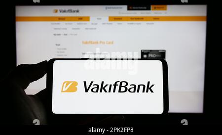 Person holding smartphone with logo of Turkiye Vakiflar Bankasi T.A.O. (VakifBank) on screen in front of website. Focus on phone display. Stock Photo