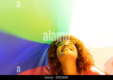 Closeup of cheerful biracial young woman wearing eyeglasses smiling under rainbow flag Stock Photo
