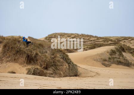 Isle of Lewis, Scotland. 5th April 2023. UK Weather: A wet and windy start to the Easter Holidays on the Isle of Lewis. A man in waterproof clothing sits in the sand dunes overlooking Eoropie Beach on an overcast and drizzly day. Credit: Bradley Taylor / Alamy Live News Stock Photo