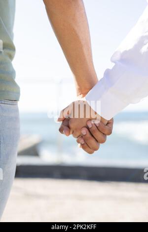 Midsection of diverse couple holding hands on promenade by the sea Stock Photo