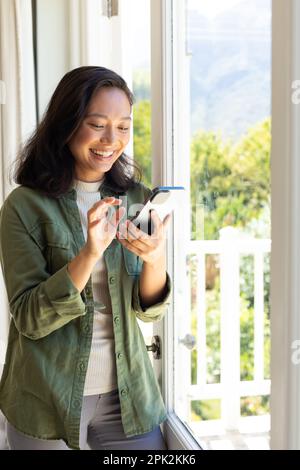 Happy asian woman standing next to window and using smartphone Stock Photo