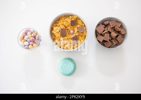 Ingredients in bowls for making chocolate nests - cornflakes, chocolate chunks, mini eggs and cupcake cases (Easter) Stock Photo