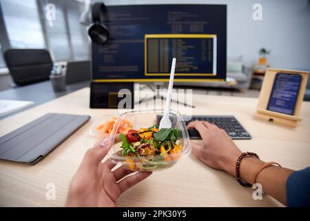 POV of unrecognizable man eating takeout lunch at workplace while programming code, copy space Stock Photo