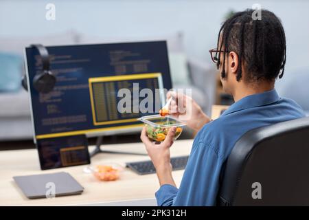 Back view of young computer programmer eating takeout lunch at workplace while writing code, copy space Stock Photo