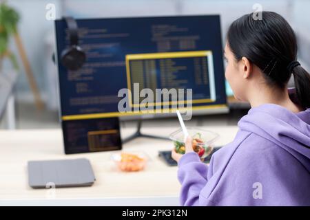 Back view of female computer programmer eating takeout lunch at workplace while writing code, copy space Stock Photo