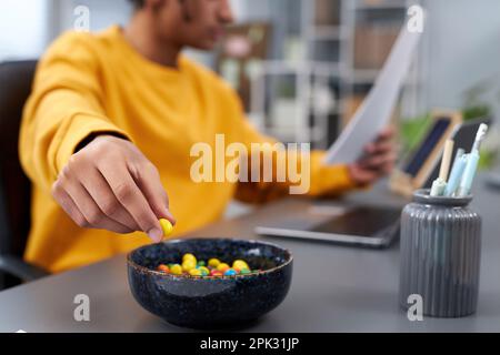Close up of young man eating snacks while working or studying at home, focus on hand holding colorful candy, copy space Stock Photo