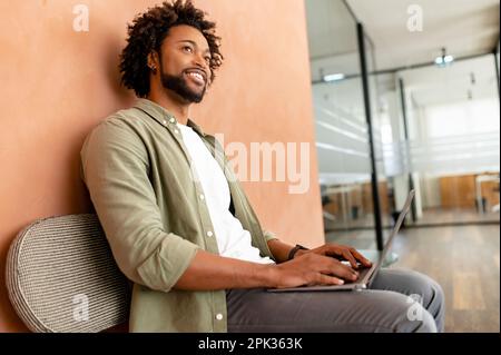 Smiling optimistic young male entrepreneur in casual wear, sitting on the chair, typing on keyboard, freelancer professional is feeling ready to work and start new projects Stock Photo