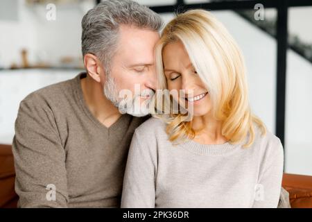Cheerful and happy middle-aged spouses enjoying time together. Handsome grey-haired man hugging gently beautiful good-looking woman, their eyes are closed. Love and bond concept. Close-up portrait Stock Photo