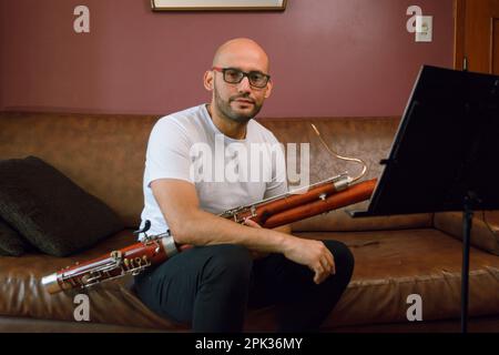 portrait of young bald latino man with glasses holding a bassoon and a music stand sitting on a brown leather sofa in the living room looking at the c Stock Photo