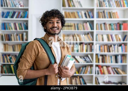 Portrait of happy satisfied african american student with curly hair, guy with books and backpack standing in university academic library smiling and looking at camera. Stock Photo