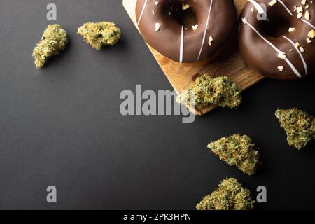 Chocolate-coated donuts with hazelnut topping lie on a wooden board among dry buds of medical marijuana.  On a black background, flatley Stock Photo