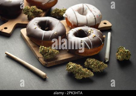 Donuts covered with chocolate and white icing lie on wooden boards among dry buds of medical marijuana, next to two joints.  On a black background Stock Photo