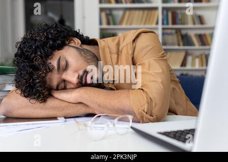 Tired student sleeping on table inside university academic library, hispanic man among books with laptop preparing for session and final exams. Stock Photo