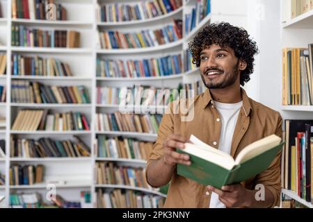 Young hispanic student reading a book while standing inside an academic university library, man smiling contentedly looking towards the window. Stock Photo