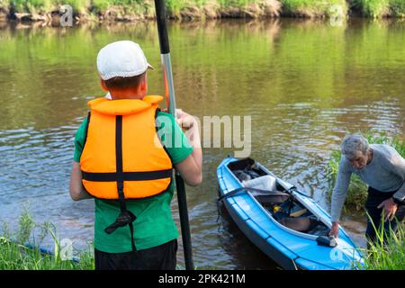 Child with a kayak paddle for rafting stands on the river bank. Family sport water hike, a summer adventure. Eco-friendly and extreme tourism, active Stock Photo