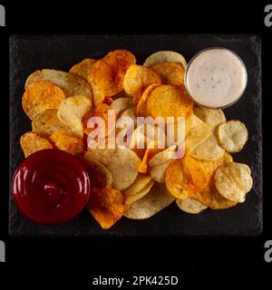 Golden brown and crunchy potato chips with zesty white and savory red dip isolated on black background Stock Photo