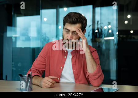 Shocked and upset young man sitting at desk in office, thoughtfully holding head with hand, closed eyes, crying, feeling pain Fired from job, salary reduction, unemployed. Stock Photo