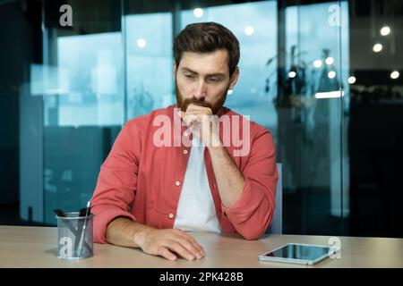 Shocked and upset young man sitting at the desk in the office, thoughtfully holding his head with his hand. Fired, downsizing, salary reduction, unemployed, company closing. Stock Photo