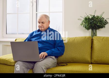 Old gray-haired senior man using laptop while sitting on couch in the living room at home. Stock Photo