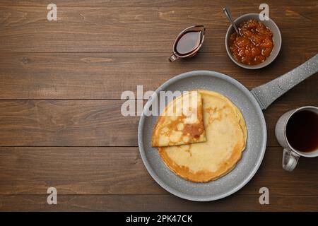 Frying pan with delicious crepes, jam, syrup and aromatic tea on wooden table, flat lay. Space for text Stock Photo