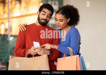 Diverse Buyers Couple Shopping Using Smartphone At Modern Mall Stock Photo