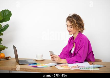Smiling Young Female Designer Using Smartphone At Workplace In Home Office Stock Photo