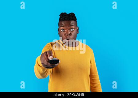 Shocked Black Man Watching Television, Pointing Remote Controller At Camera Stock Photo