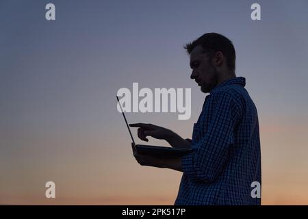 Hardly working evening: male freelancer developer or it support manager in silhouette working outdoor using laptop at sunset. Adorable dusk at background. High quality image Stock Photo