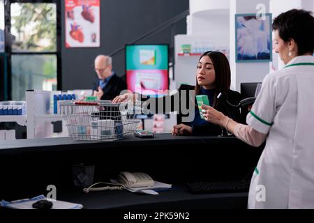 Pharmacy buyer giving cashier medications for scanning at drugstore counter desk. Pharmaceutical employee standing at checkout and using scanner on vitamin packages barcode Stock Photo