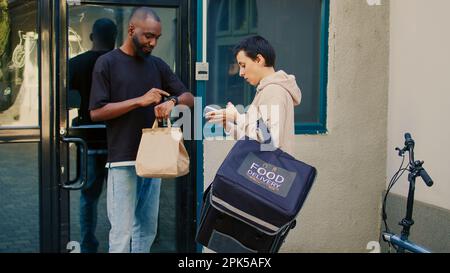 Male customer taking restaurant food order using smartwatch to pay with nfc on pos, paying for takeaway lunch. Food delivery service courier giving fastfood in paperbag, contactless terminal. Stock Photo